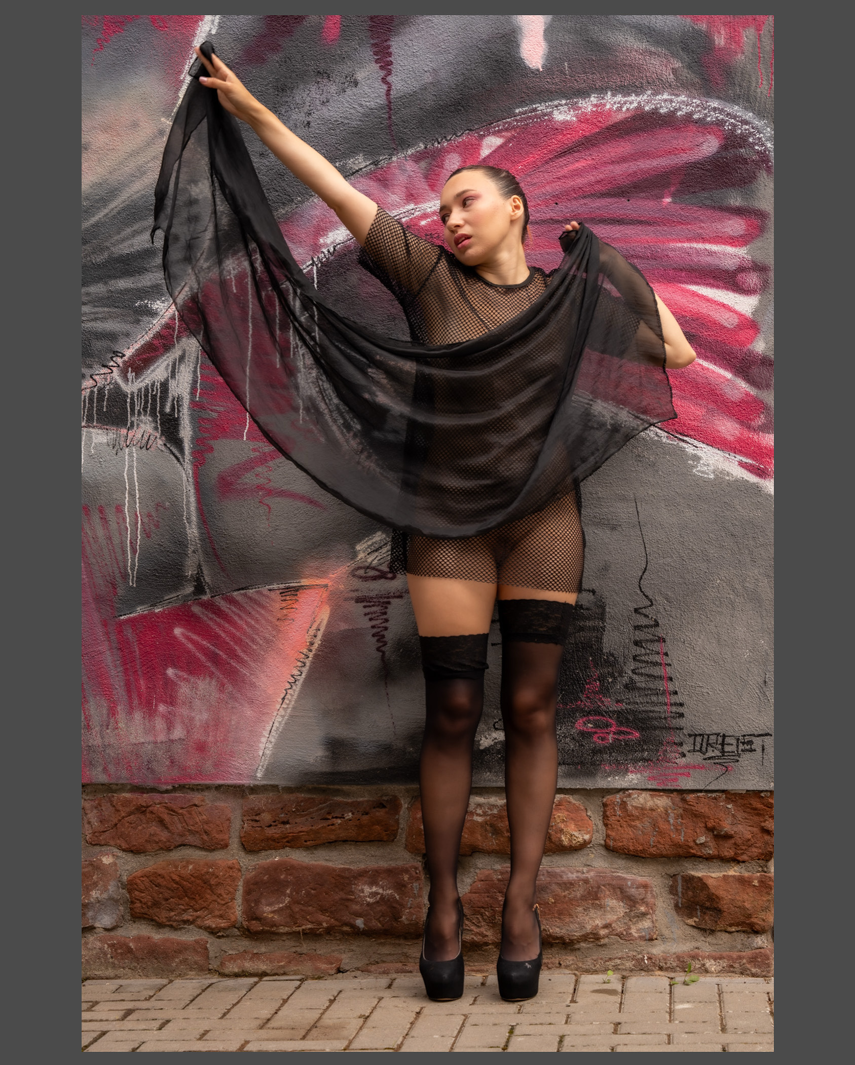 Junge Frau trägt ein schwarzes Netzoutfit, halterlose schwarze Strümpfe und schwarze High Heels und posiert mit einem schwarzen Tuch vor einer bunten Graffiti-Wand