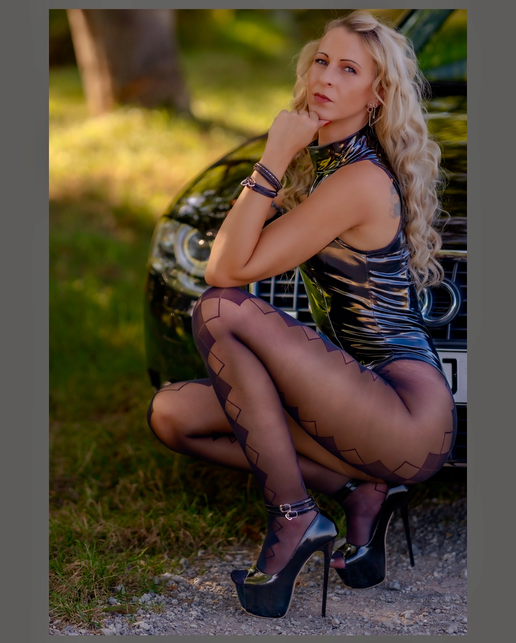 Franzi kniet vor ihrem Auto in einem Latexoutfit und Strumpfhosen mit einem geometrischen Muster, ihre Beine sind lang wie der Montag Morgen
