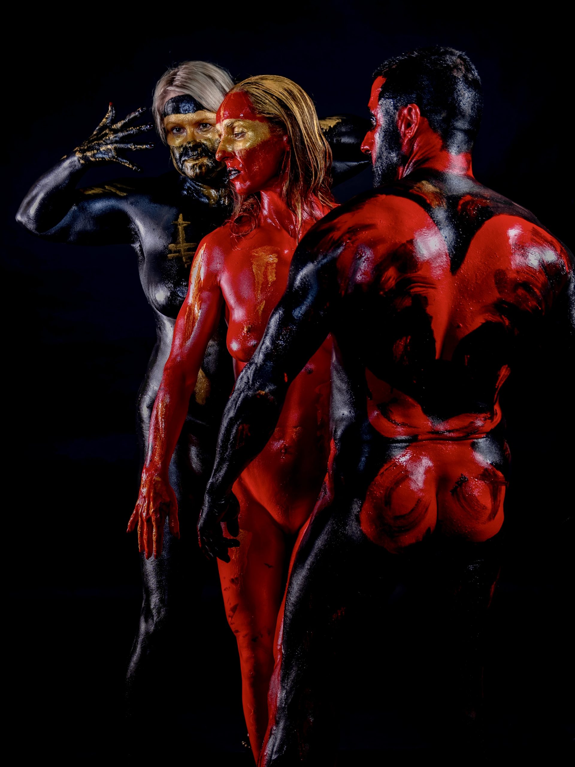 Drei Körper in auffälligem Bodypainting aus Rot, Schwarz und Gold posieren kunstvoll auf dunklem Hintergrund