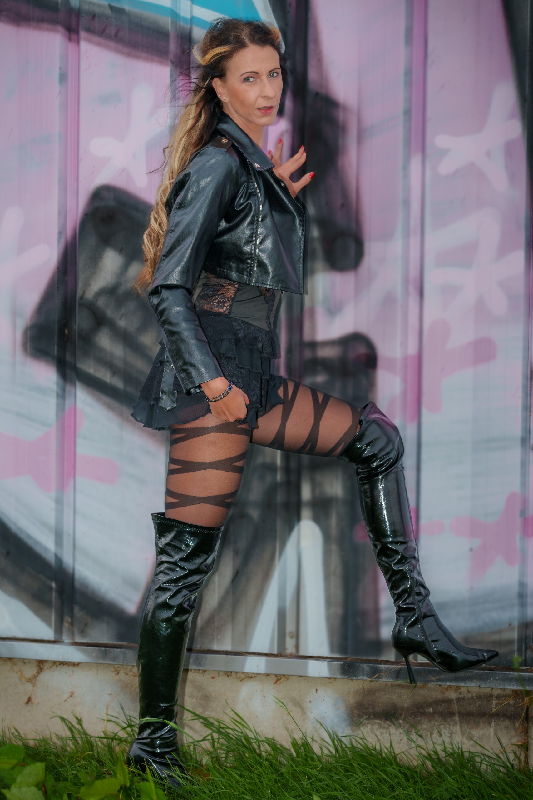 Schlanke Frau in Stiefeln, geometrische gemusterte Strumpfhose und Lederjacke vor einer Wand