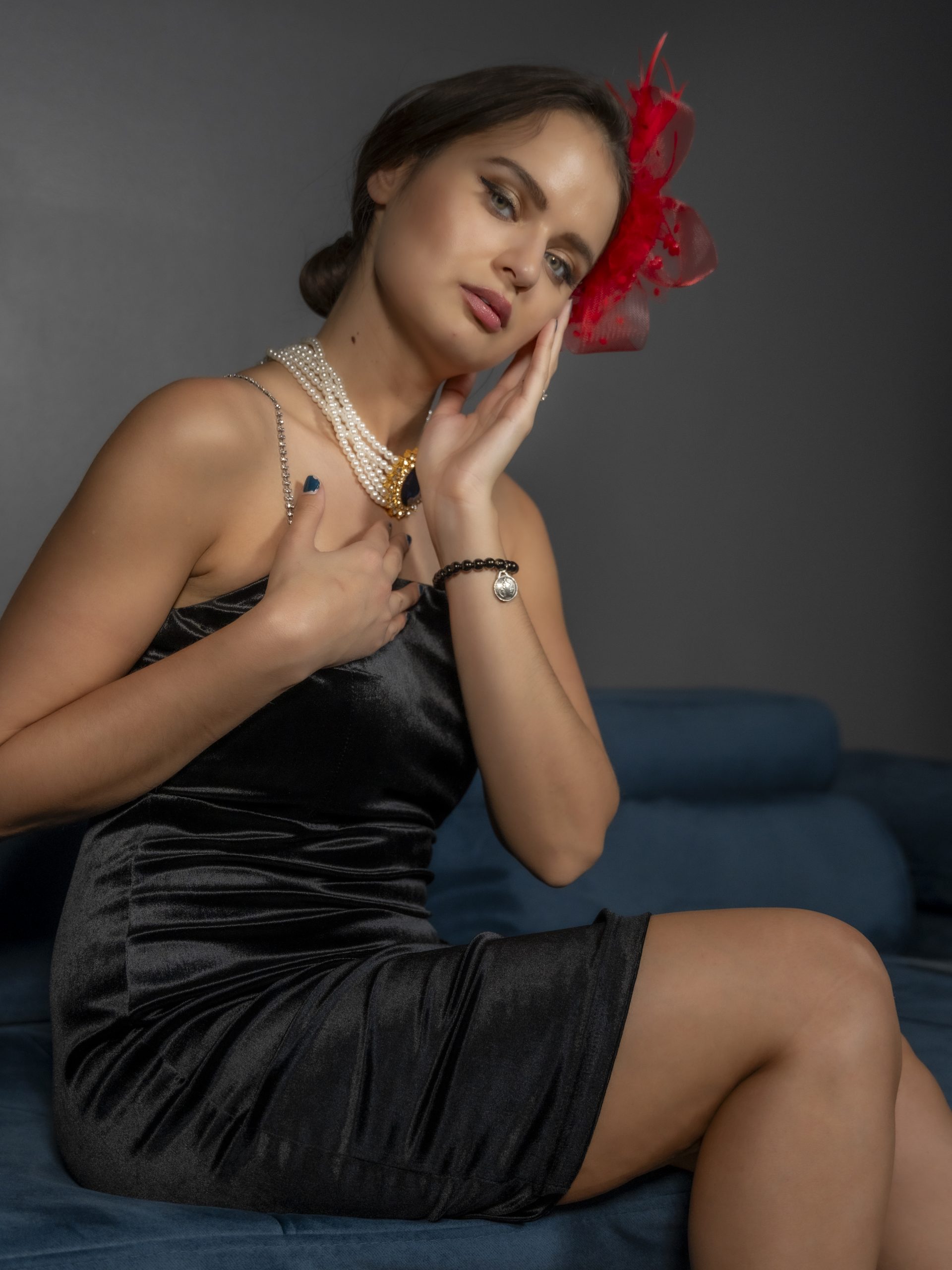 Frau im schwarzen Samtkleid mit Perlenkette und rotem Fascinator posiert stilvoll auf einem blauen Sofa