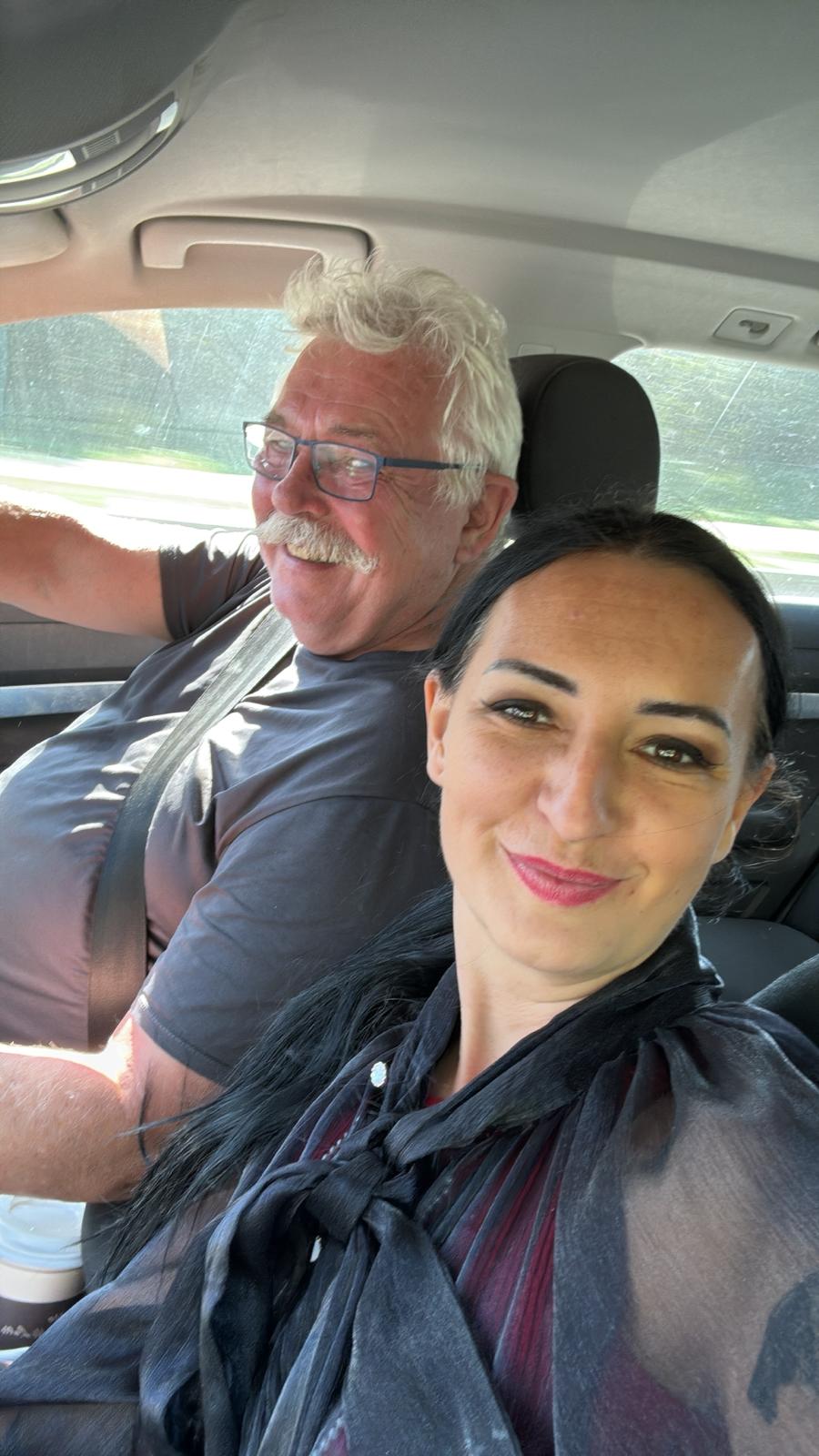 Selfie von zwei Personen im Auto, beide lächeln fröhlich während der Fahrt bei Sonnenschein