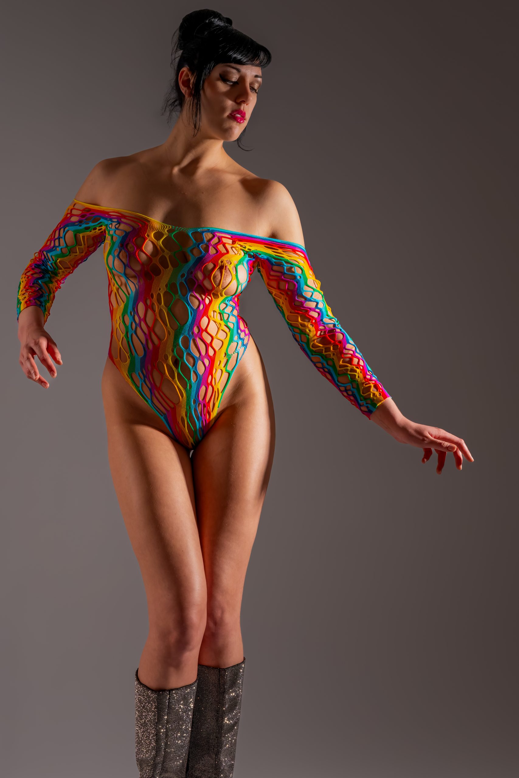 Eine Frau steht vor einem neutralen grauen Studiohintergrund. Sie trägt ein farbenfrohes, netzartiges Body-Outfit mit langen Ärmeln und glitzernde Stiefel. Ihr dunkles Haar ist hochgesteckt, der Blick ist nach unten gerichtet. Das Bild wirkt modisch, dynamisch und elegant.