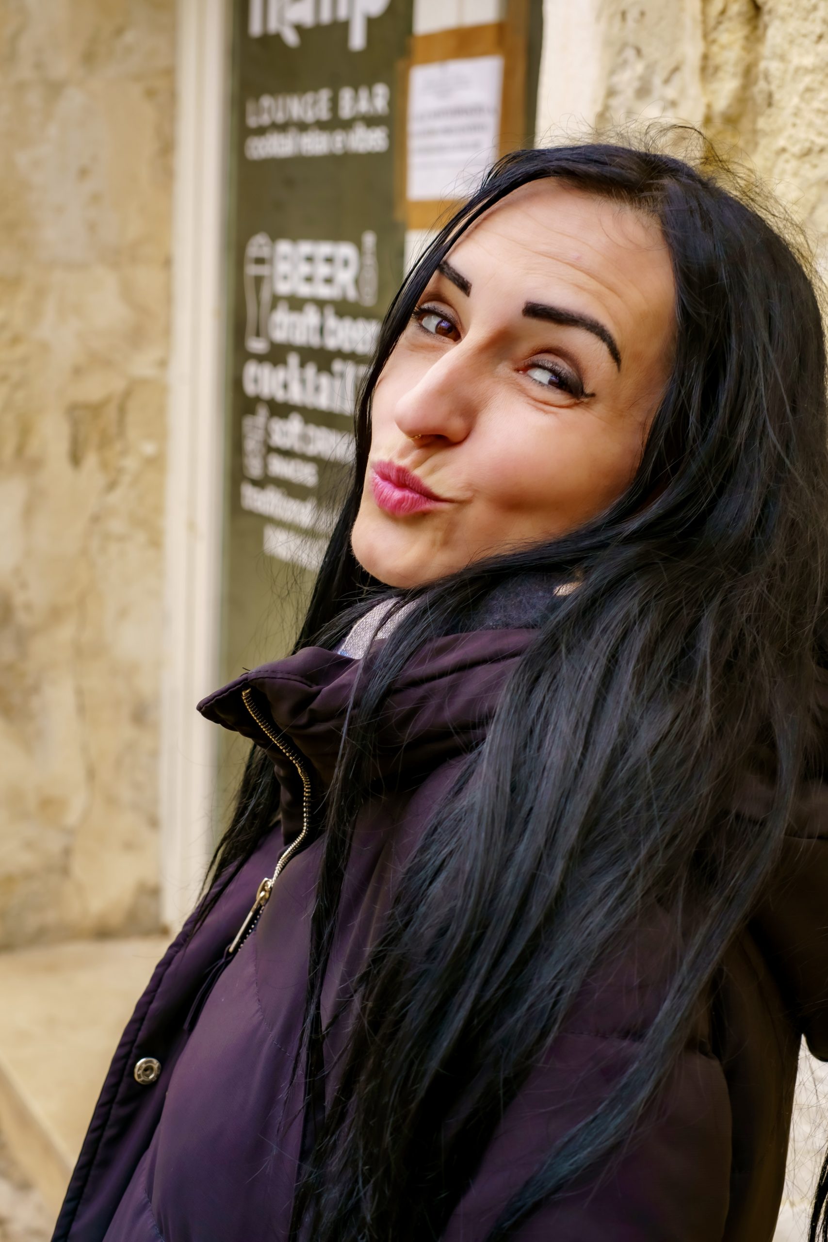 Kira mit langen dunklen Haaren trägt eine dunkle Jacke, blickt seitlich in die Kamera und macht eine verspielte Kussmund-Geste vor einem unscharfen urbanen Hintergrund.
