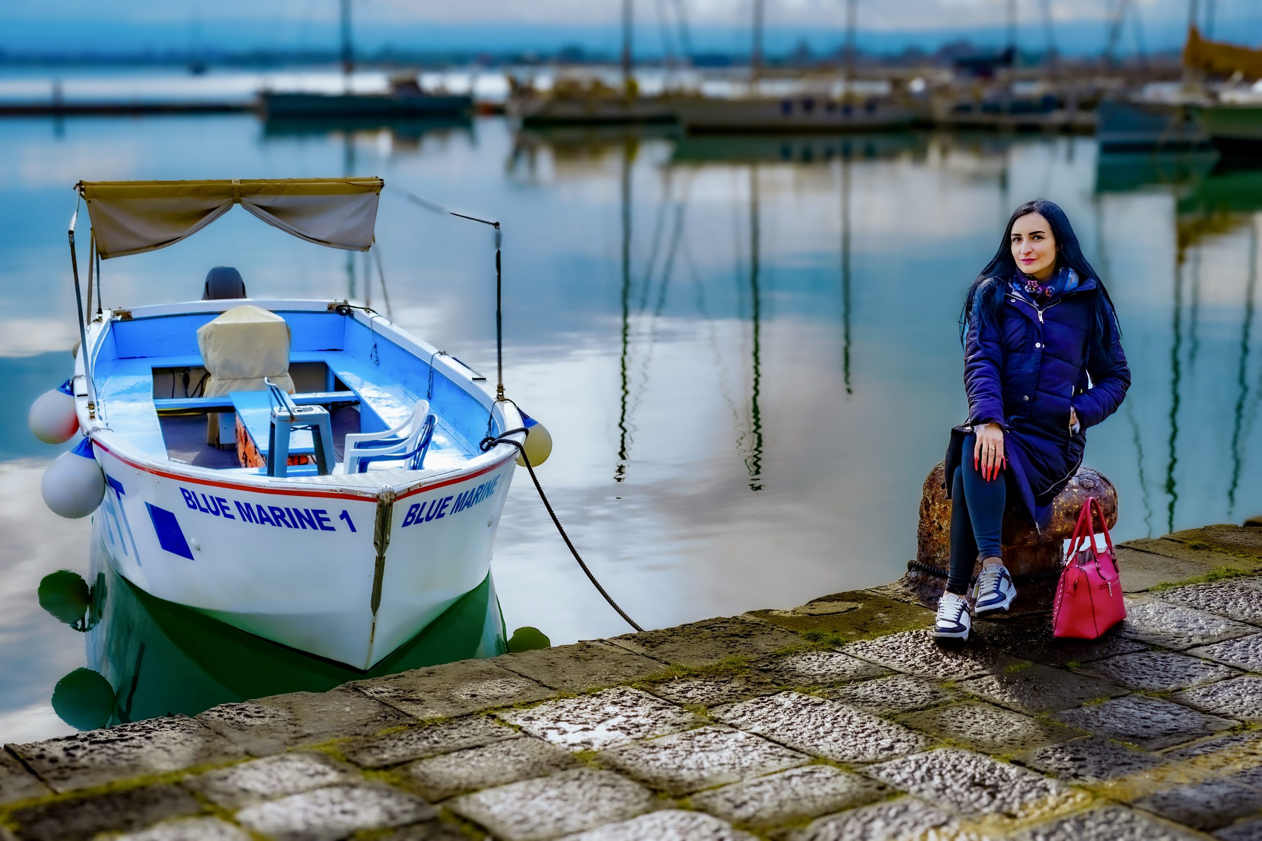 Kira sitzt in dunkler Jacke und sportlichem Outfit am Hafen von Syrakus neben einer roten Tasche, links liegt ein weiß-blaues Boot im ruhigen Wasser.
