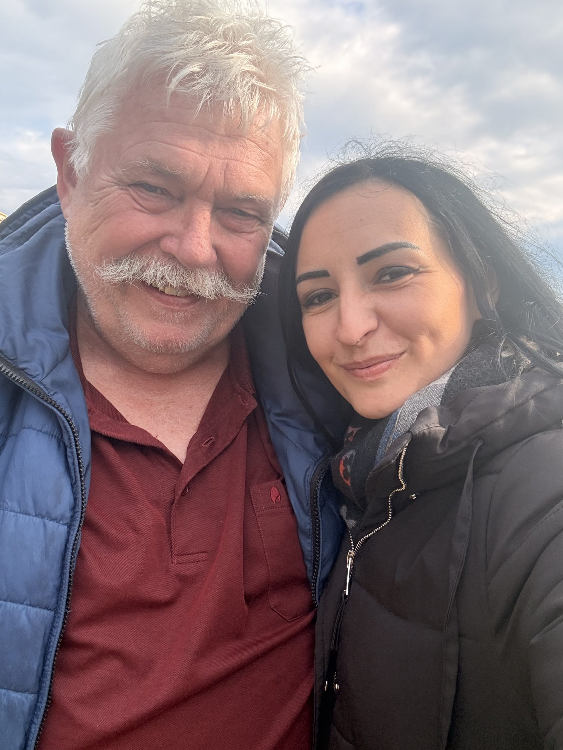 „Henry und Kira machen ein Selfie im Urlaub auf Sizilien, beide lächeln in die Kamera vor hellem, bewölktem Himmel.“