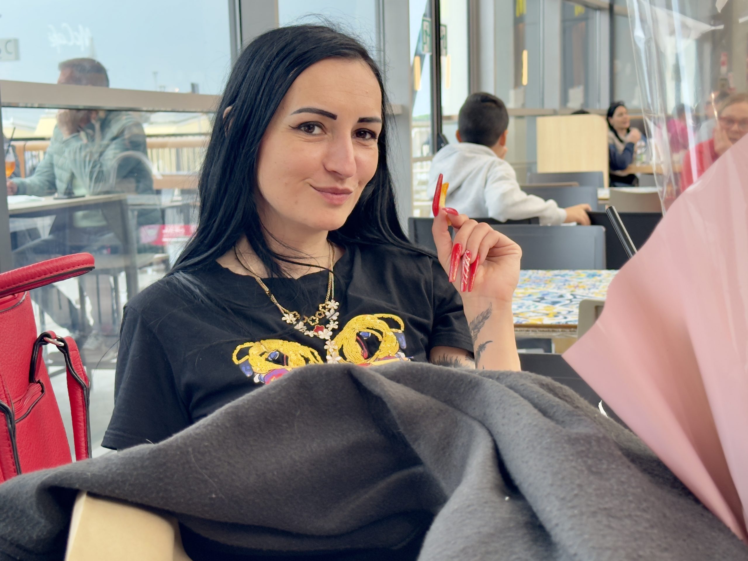 Porträt einer Person mit schwarzen Haaren, schwarzem T-Shirt, roter Handtasche und auffälligen roten Nägeln, sitzend in einem hellen Café während eines gemeinsamen Bummels in Syracusa.