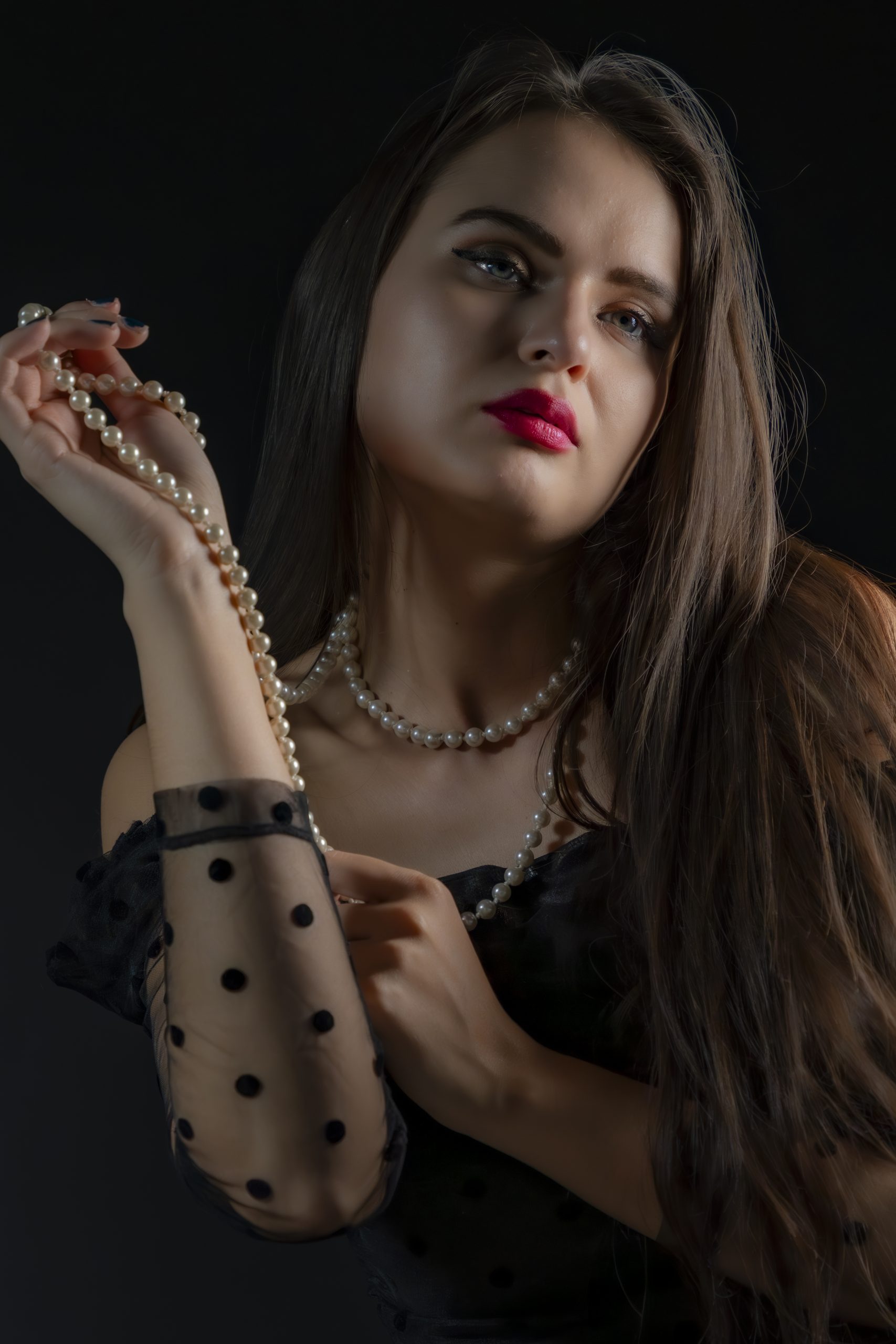 Portrait einer jungen Frau vor dunklem Hintergrund. Sie trägt ein schwarzes Outfit mit transparentem, gepunktetem Ärmel und hält eine Perlenkette in der Hand. Eine weitere Perlenkette liegt um ihren Hals. Ihr langes braunes Haar fällt über die Schulter, sie blickt mit ruhigem, intensivem Ausdruck in die Kamera. Roter Lippenstift und weiches Seitenlicht setzen elegante Akzente.