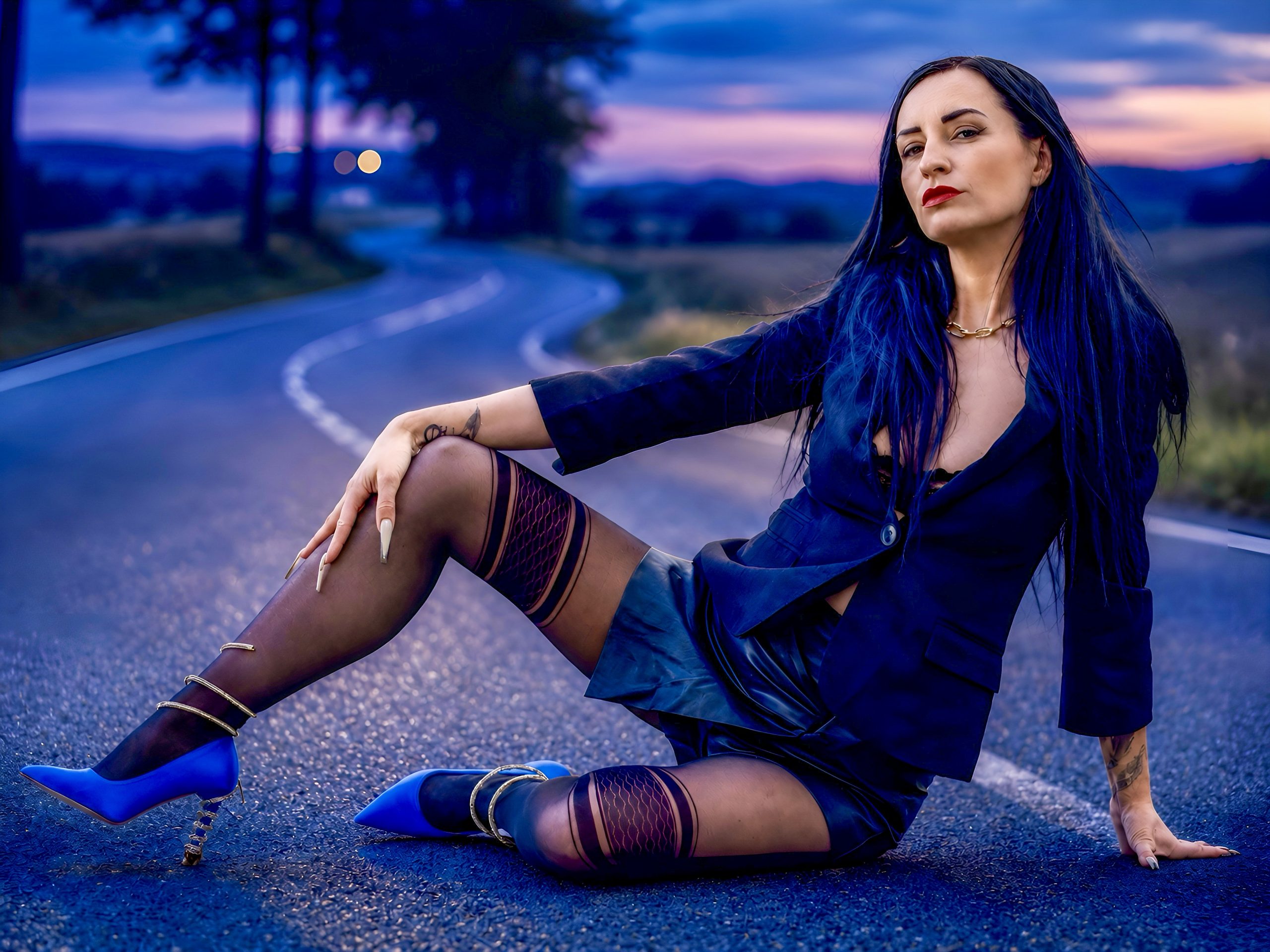 Kira in dunklem Fashion-Outfit mit Blazer, Lederrock, gemusterter Strumpfhose und blauen High Heels sitzt selbstbewusst auf einer Straße in der blauen Stunde.