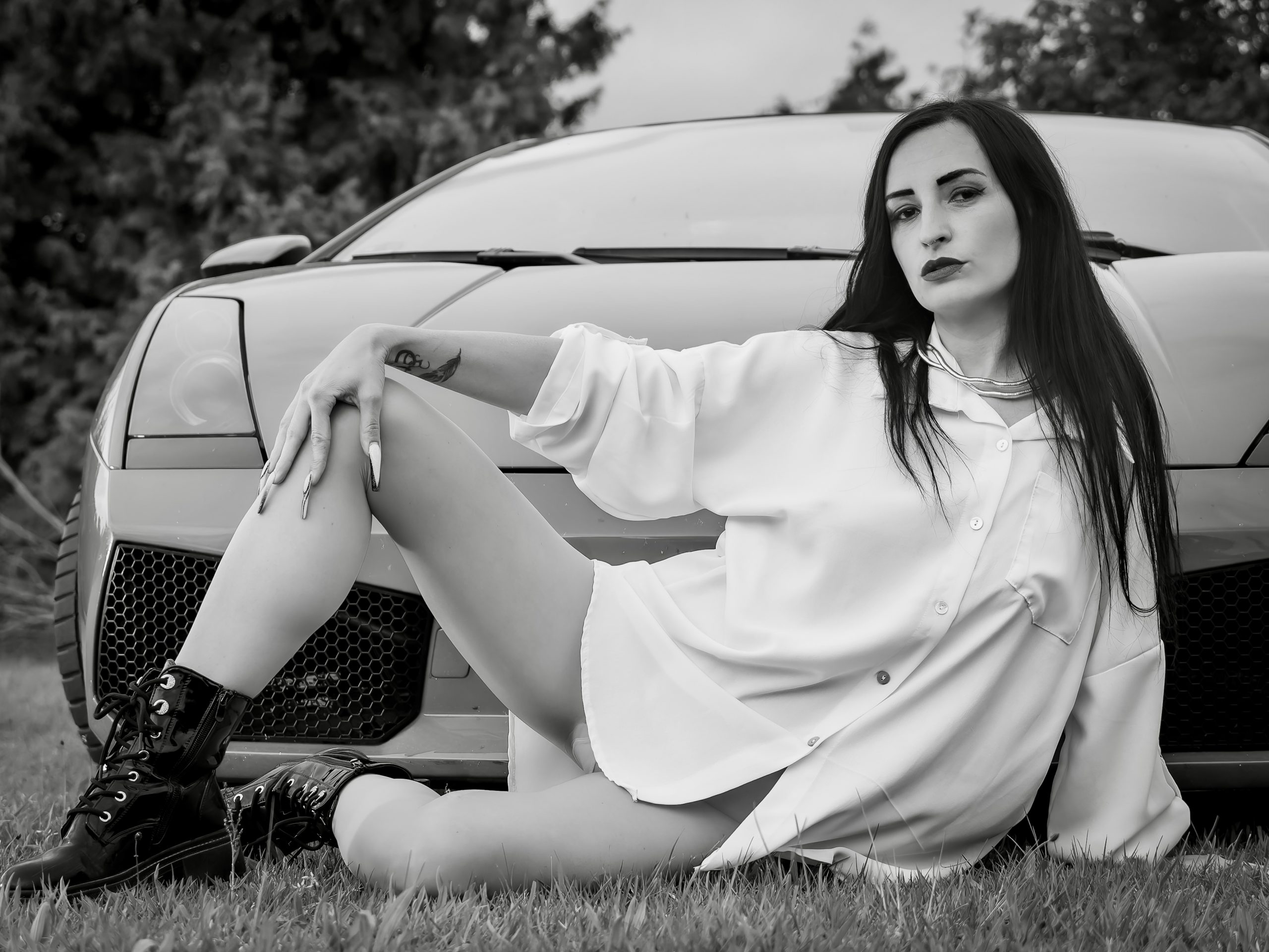 Frau mit langen dunklen Haaren sitzt in weißem Oversize-Hemd, feiner Strumpfhose und schwarzen Boots vor einem Sportwagen auf einer Wiese; das Bild ist in Schwarzweiß gehalten.