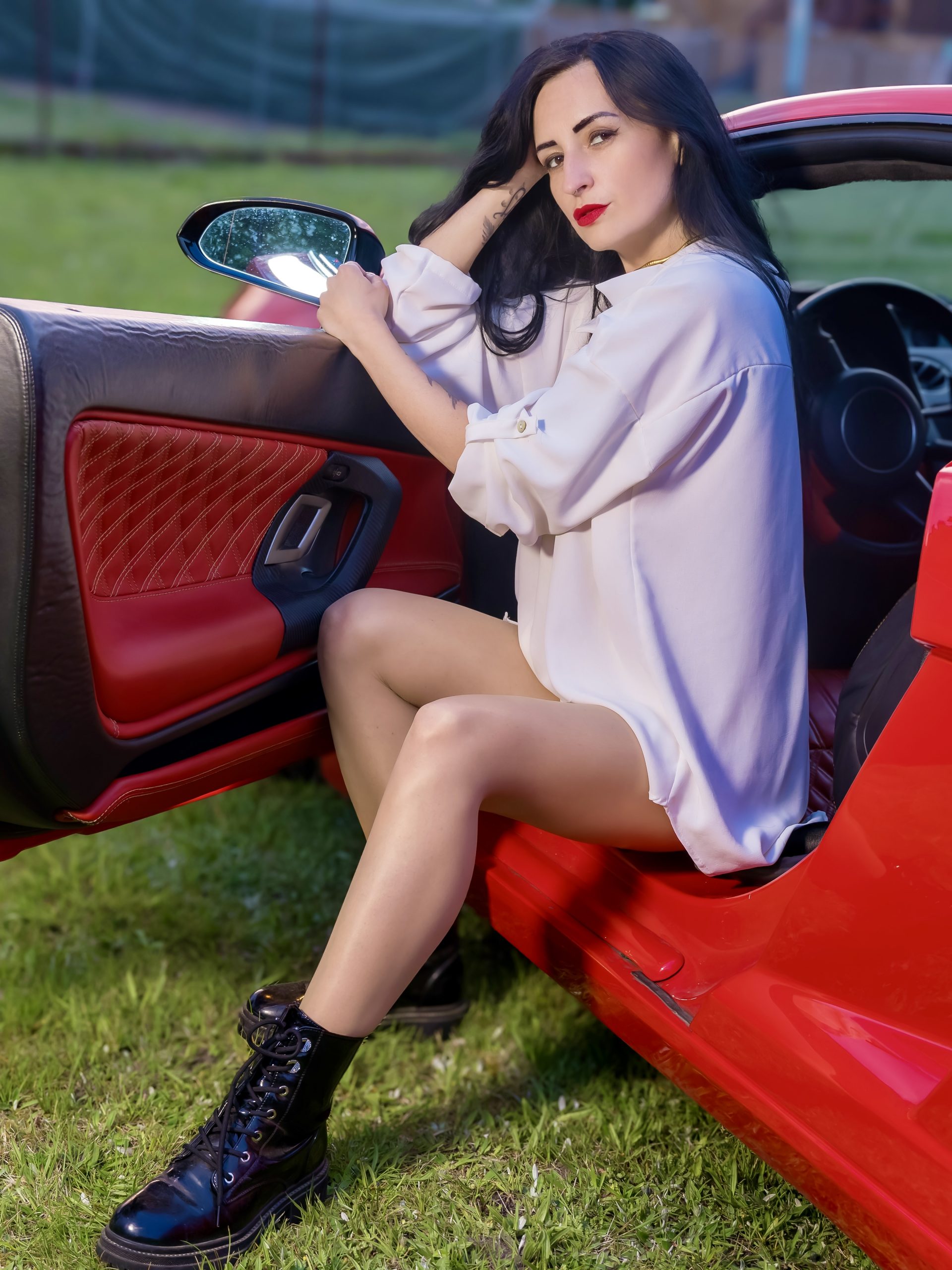 Porträt von Kira in einem roten Lamborghini auf einer Wiese. Sie sitzt bei geöffneter Tür im Wagen, trägt ein helles feminines Outfit, schwarze Stiefel und Falke seidenglatt Strumpfhose und schaut direkt zur Kamera.