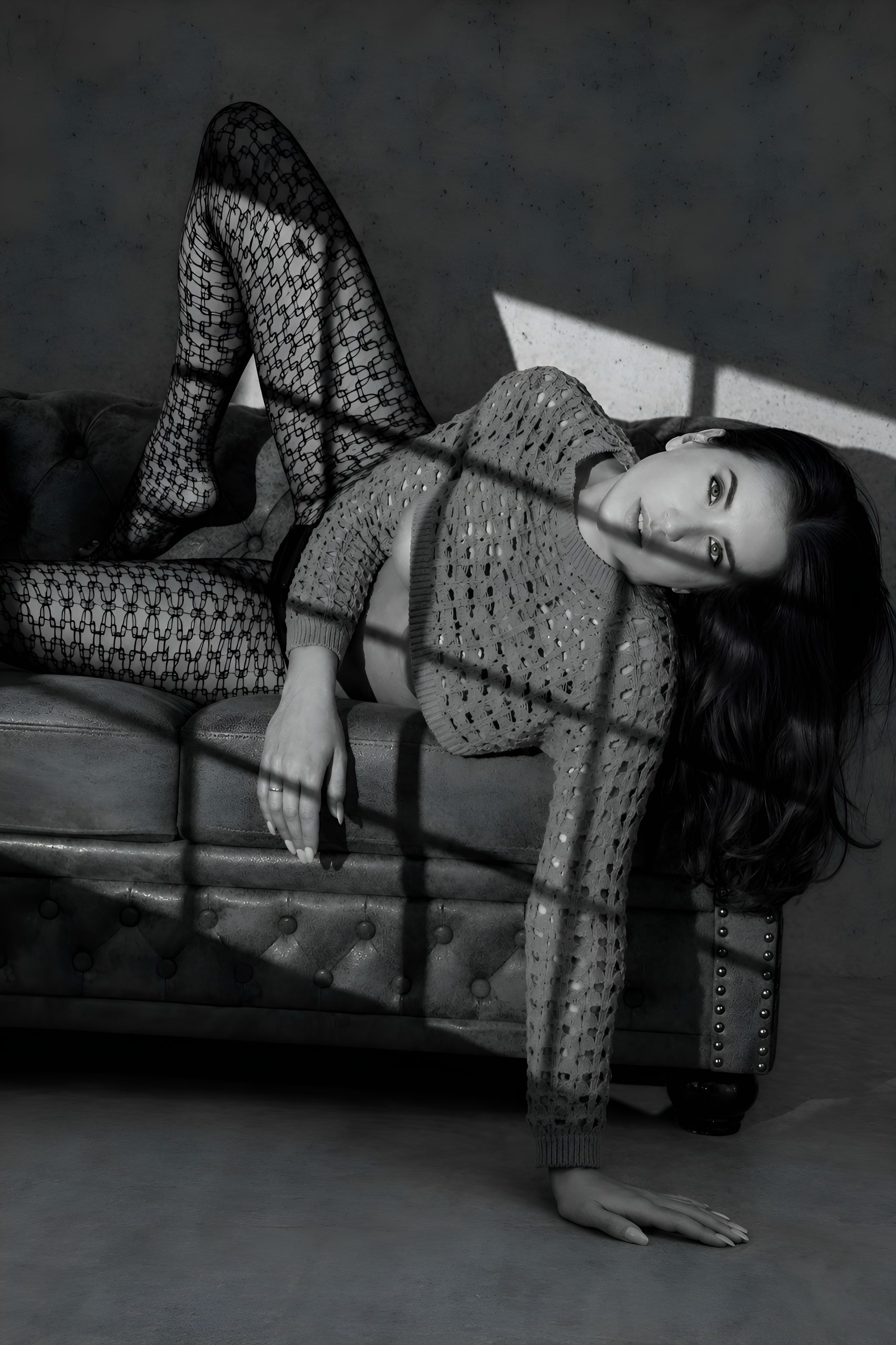 Model Sabrina liegt auf einer gepolsterten Couch in dramatischem Schwarz-Weiß-Licht. Sie trägt einen strukturierten Strickpullover und gemusterte Strumpfhosen, während harte Schatten im Newtonstyle eine provokante, elegante Fashion-Stimmung erzeugen.