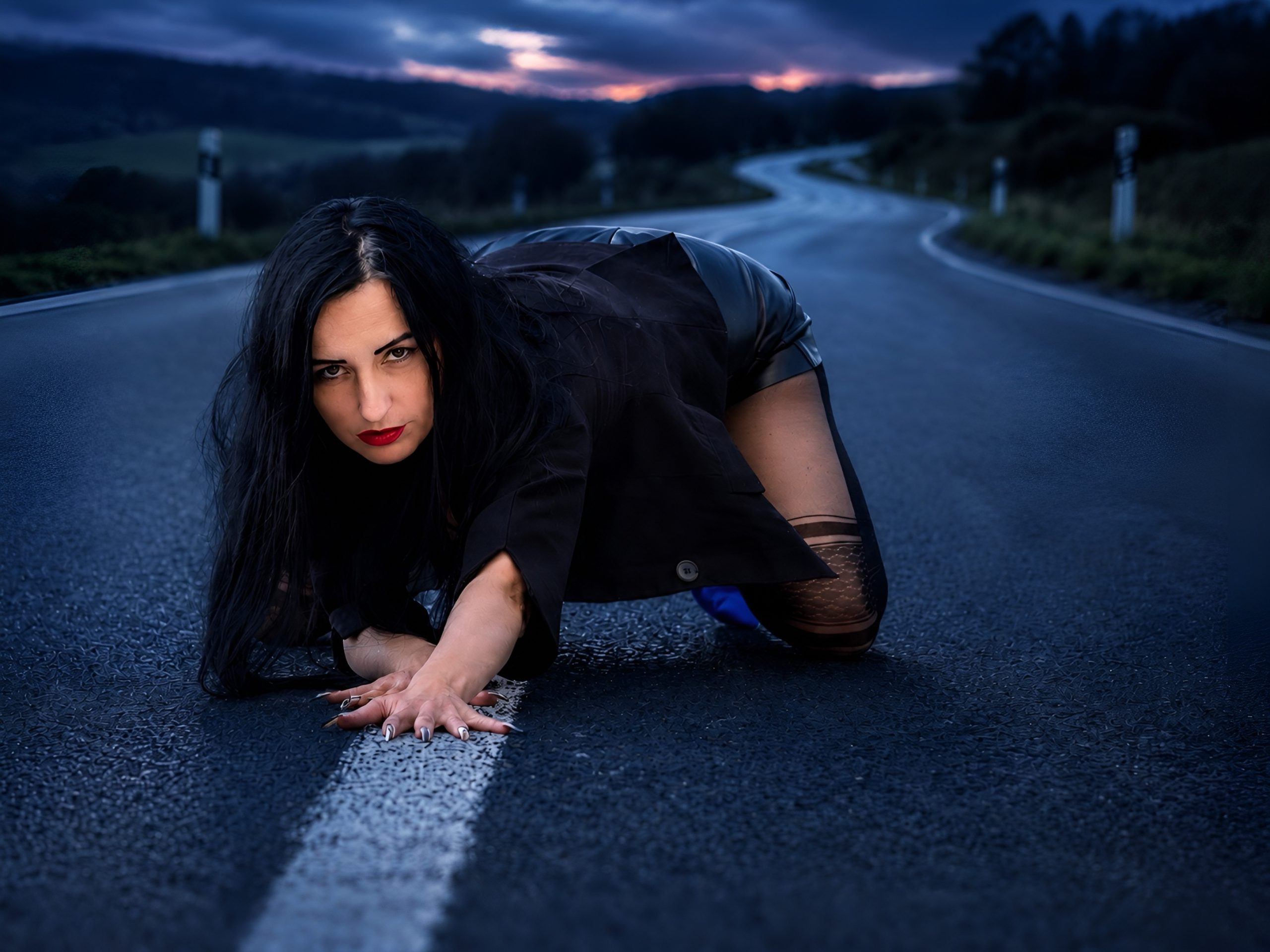 Frau mit langen dunklen Haaren in Blazer, kurzem Rock und feinen gemusterten Nylons kniet bei Dämmerung auf einer leeren Straße und blickt intensiv in die Kamera.