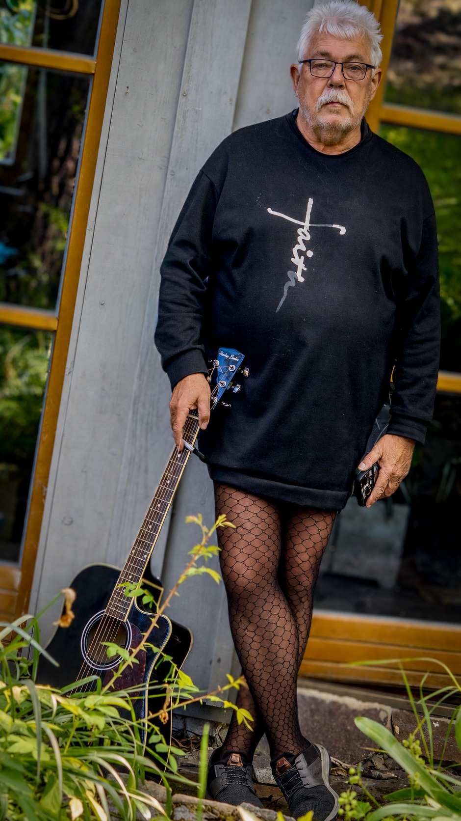 Henry steht mit Gitarre vor einer Garten- und Holzfassadenkulisse. Sein schwarzes Outfit mit markantem Muster, der direkte Blick und die ruhige Haltung zeigen eine mutige, künstlerische Selbstinszenierung.