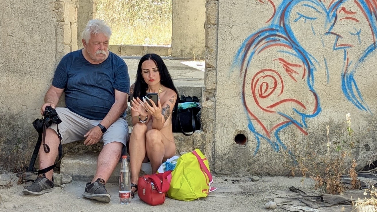 Fotograf Henry und Model Kira sitzen während einer kleinen Shootingpause in einer verlassenen Location neben Kamera, Taschen und Wasserflasche vor einer Wand mit farbigem Graffiti.