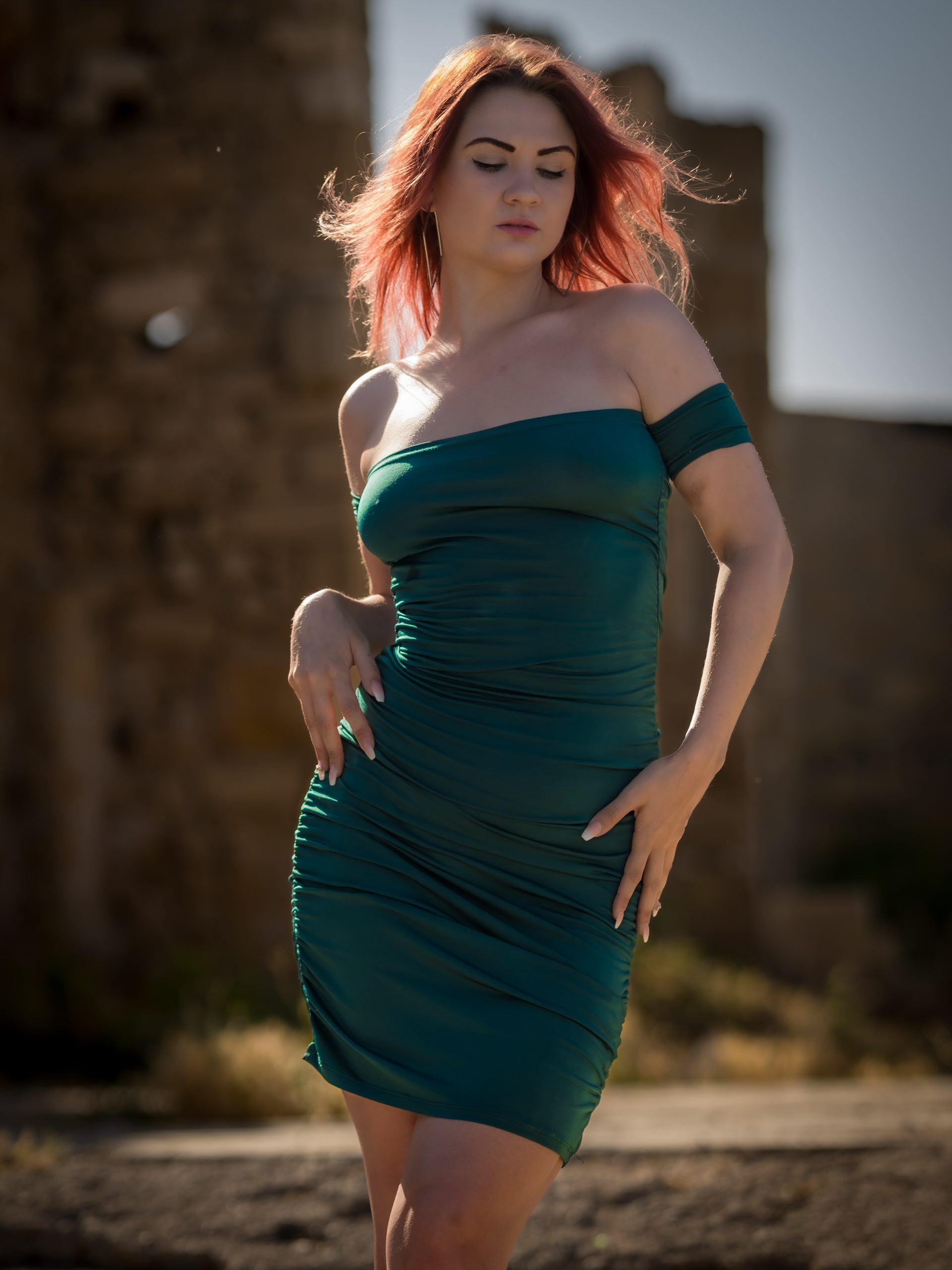 Kamila steht im Freien vor einer weich unscharfen Ruinenkulisse und trägt ein eng anliegendes grünes schulterfreies Kleid. Ihr rötliches Haar wird vom Licht betont, während sie den Blick nach unten richtet und elegant posiert.