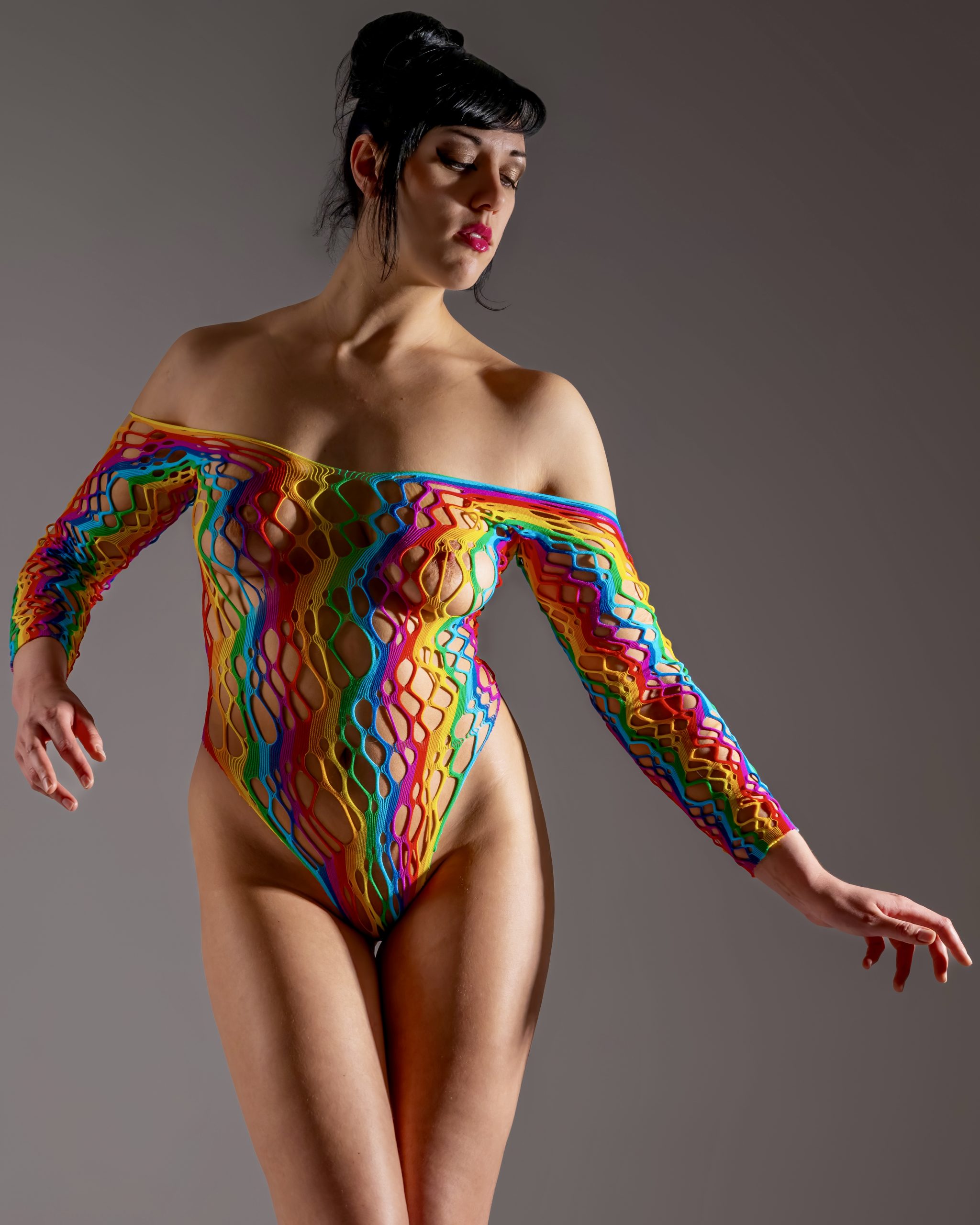 Hanna posiert vor grauem Studiohintergrund in einem farbintensiven, netzartigen Body mit gesenktem Blick und eleganter Armhaltung.