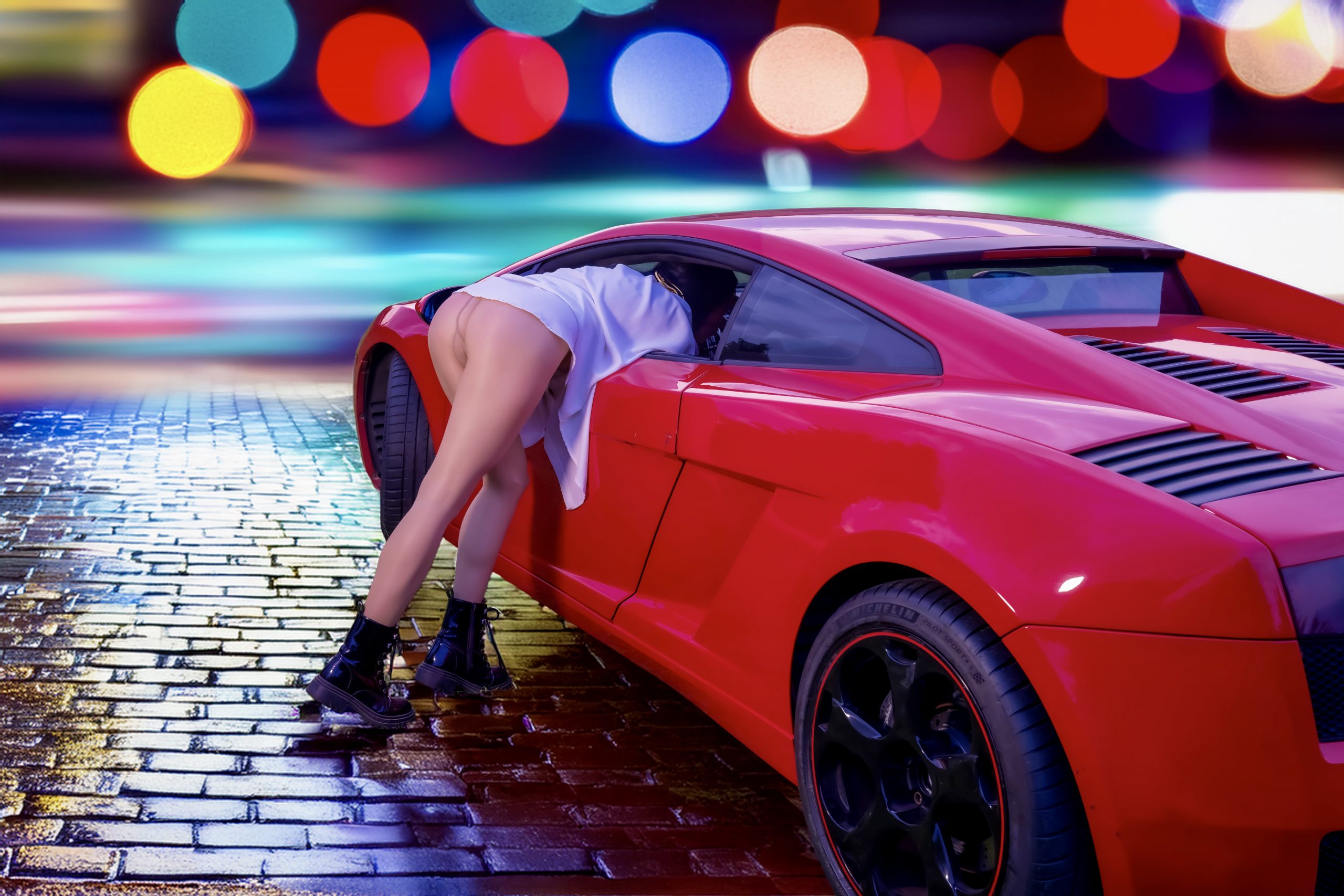 Kira lehnt sich bei Nacht in einen roten Lamborghini. Sie trägt ein oversized weißes Hemd, feine Strumpfhosen und klobige schwarze Schnürboots. Das nasse Kopfsteinpflaster spiegelt bunte Neonlichter und erzeugt eine urbane, filmische City-Atmosphäre.