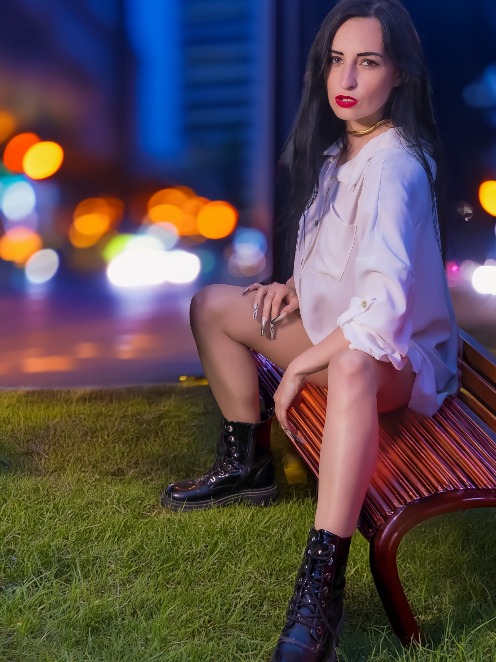 Kira sitzt nachts auf einer Bank, trägt ein oversized weißes Hemd, feine Strumpfhosen, klobige schwarze Boots und einen goldenen Halsreif vor unscharfen City-Lichtern.
