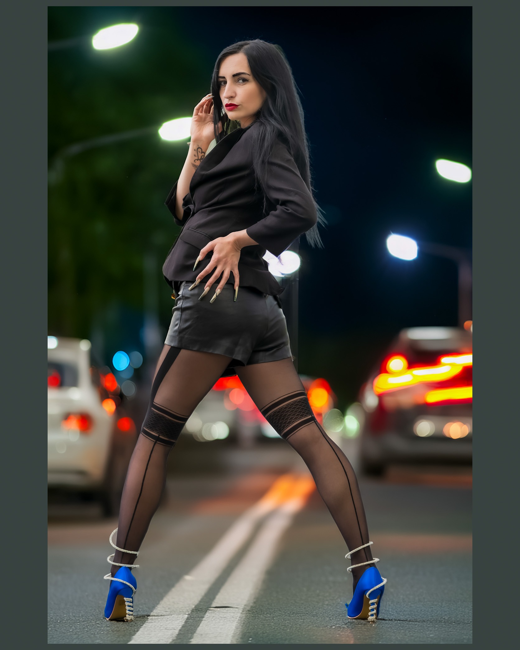Kira posiert nachts auf einer Straße vor unscharfen Autolichtern. Sie trägt ein schwarzes Outfit mit blauen High Heels und zeigt eine selbstbewusste, urbane Fashion-Pose.