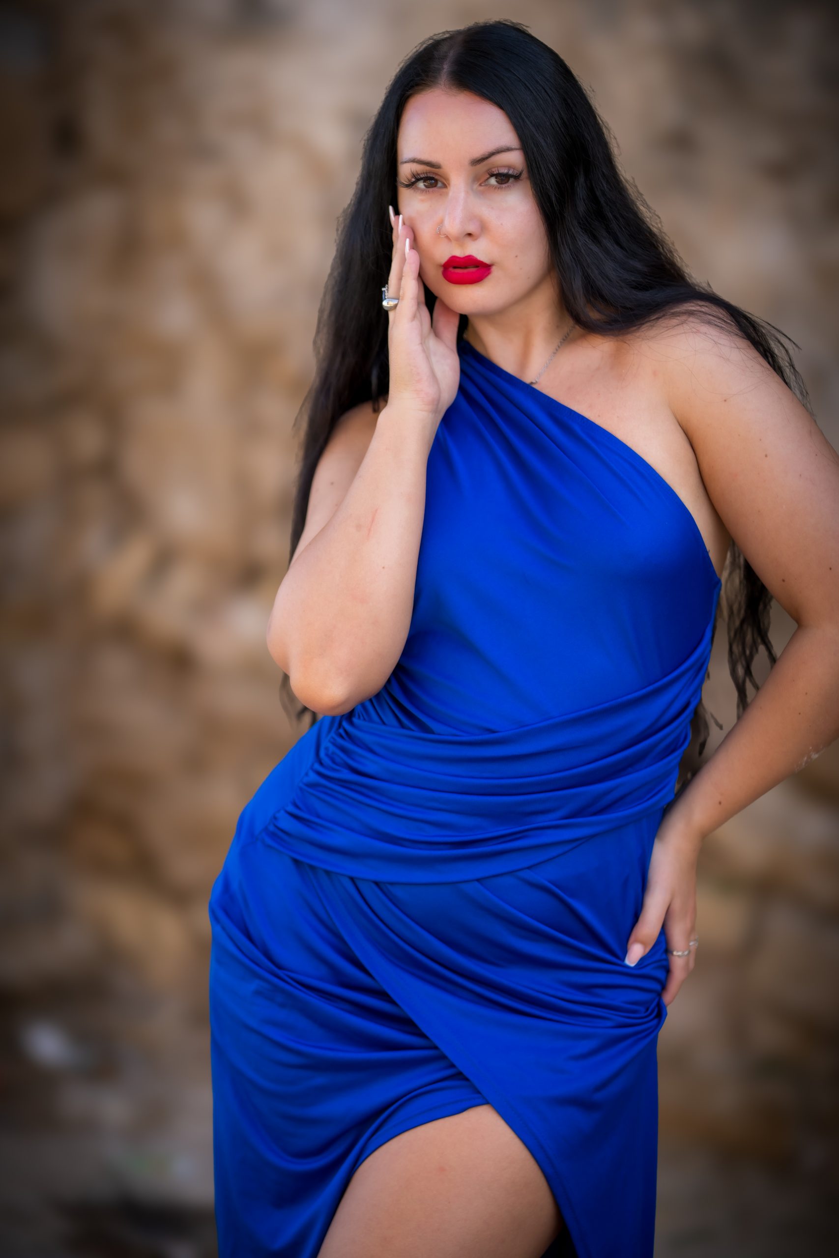 Porträt einer Frau mit langen dunklen Haaren und rotem Lippenstift in einem leuchtend blauen One-Shoulder-Kleid vor unscharfem, natürlichem Felsenhintergrund.
