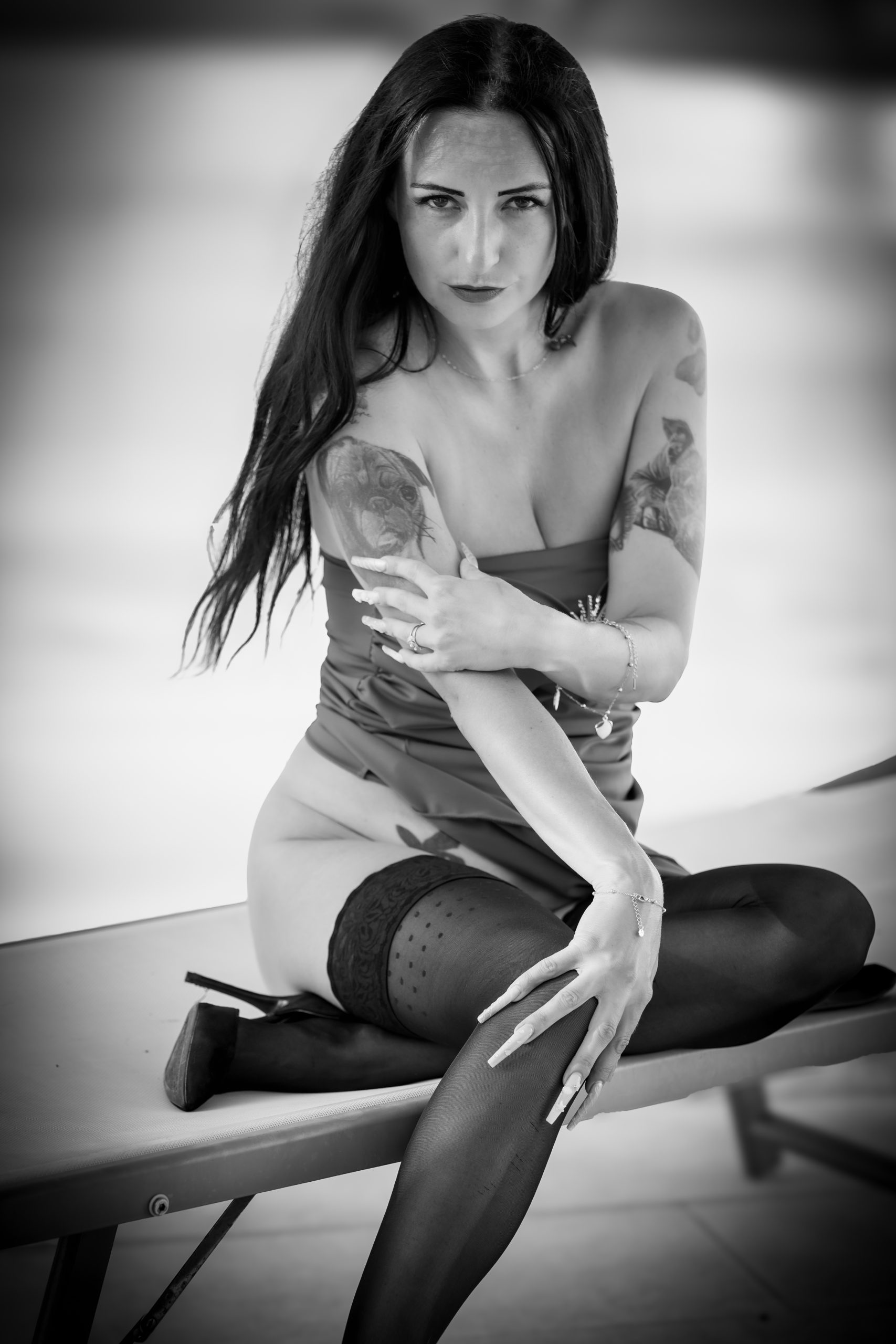 Schwarz-Weiß-Porträt einer Frau mit langen dunklen Haaren, Tattoos, elegantem Outfit und schwarzen Strümpfen, sitzend auf einer Liege mit direktem Blick in die Kamera.