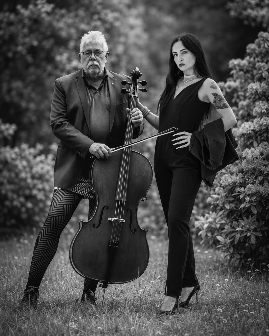 Schwarzweiß-Fashionfotografie eines Mannes und einer dunkelhaarigen Frau mit einem Cello im Freien. Der Mann trägt Sakko und eine geometrisch gemusterte Strumpfhose, die Frau einen eleganten schwarzen Jumpsuit und High Heels. Das Cello steht zentral zwischen beiden Personen. Henry Freisinger Fotografie, Fashionfotografie, Schwarzweißfotografie, Cello Fotografie, Musiker Porträt, Fashion Editorial, gemusterte Strumpfhose, Tights Fashion, außergewöhnliche Fashionfotografie, inszenierte Fotografie, Musik und Mode, Peoplefotografie, kreative Porträtfotografie