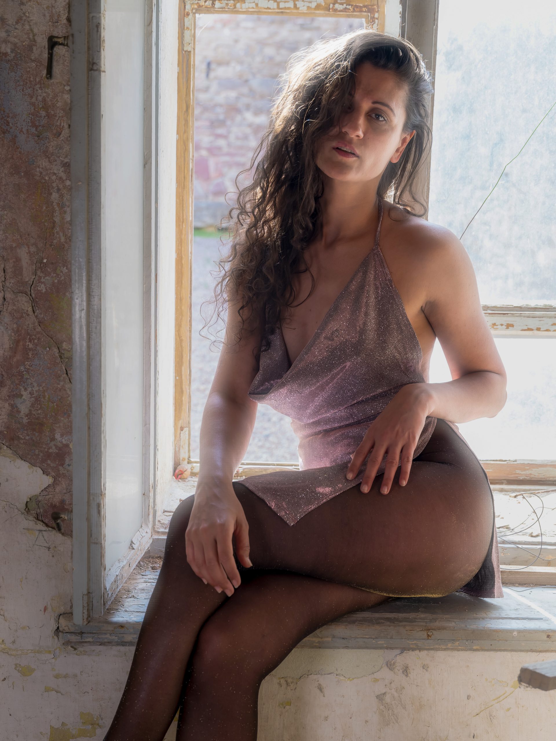 Frau mit langen dunklen Haaren in einem schimmernden roséfarbenen Kleid und dunkler Feinstrumpfhose, sitzend auf einer alten Fensterbank in einem verlassen wirkenden Raum mit starkem natürlichem Gegenlicht.