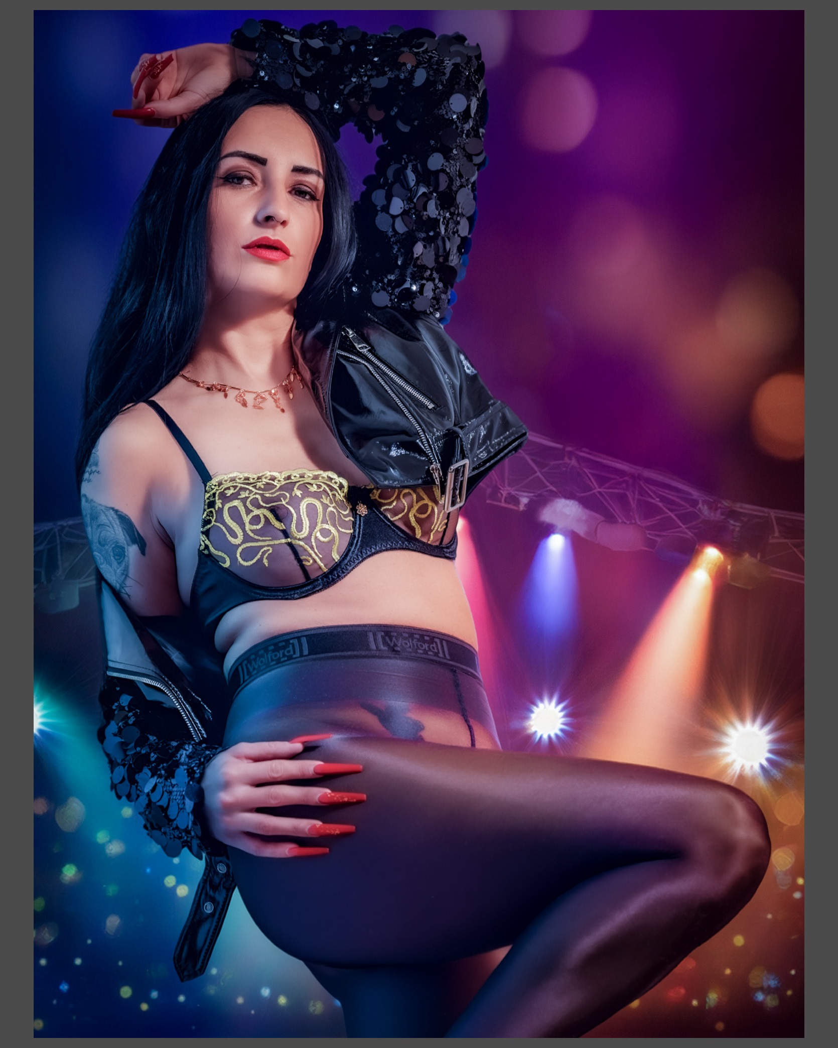 Fashionaufnahme einer Frau in schwarzer Wolford Neon 40 Feinstrumpfhose, schwarzem Body mit goldener Stickerei und glänzender Lederjacke vor einer intensiv blau, magenta und warm beleuchteten Kulisse.