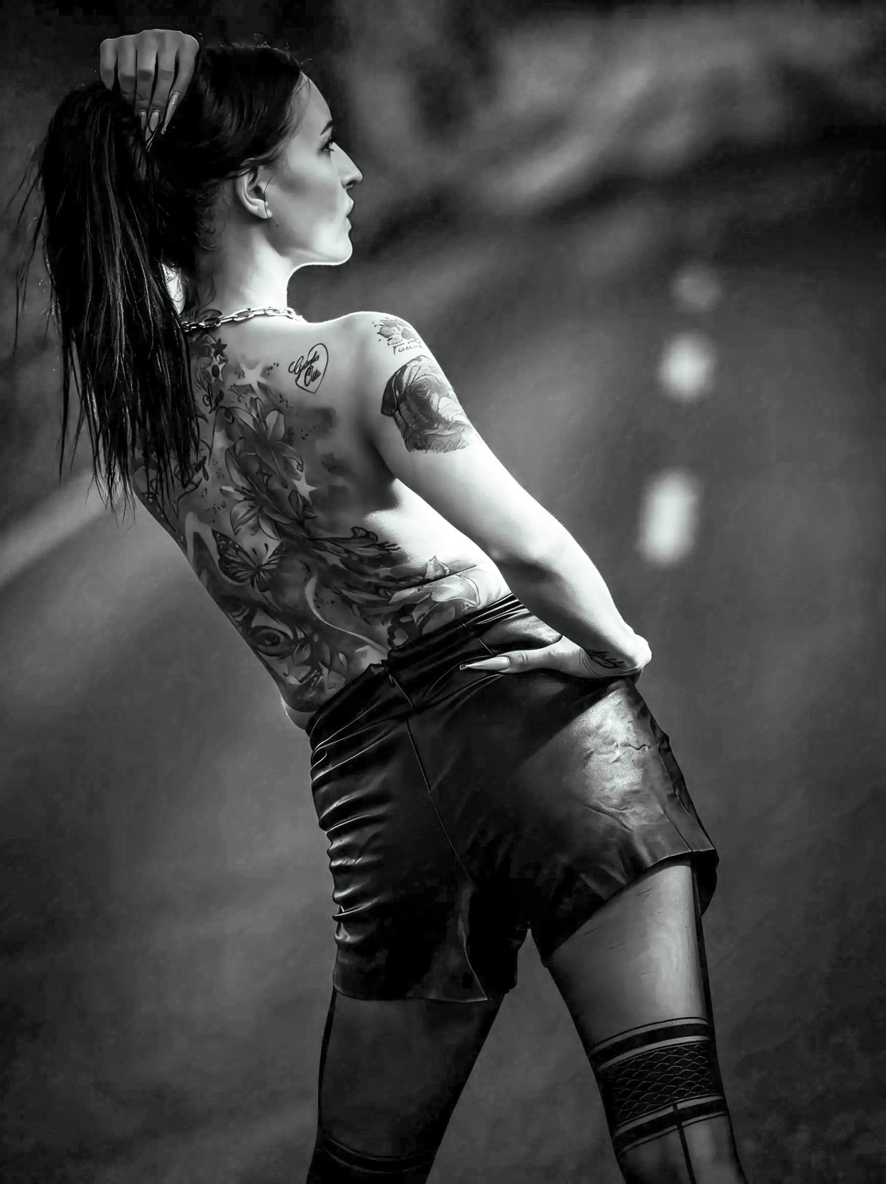 Schwarzweiß-Fashionaufnahme von Kira auf einer Straße mit ausgeprägter Perspektive. Sie trägt schwarze Ledershorts und eine dunkle gemusterte Strumpfhose. Großflächige Tattoos, Pose und Straßenflucht bestimmen die grafische Bildwirkung.