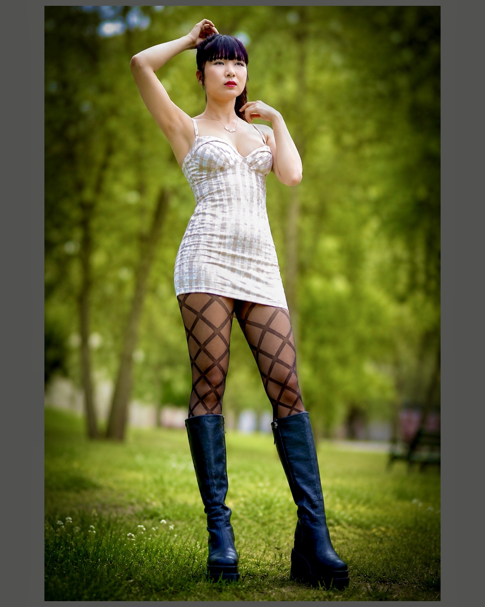 Dunkelhaarige Frau steht in einem grünen Park und blickt leicht seitlich an der Kamera vorbei. Sie trägt ein kurzes, helles Kleid mit feinem Karomuster, schwarze kniehohe Stiefel und eine transparente schwarze Feinstrumpfhose mit großem geometrischem Rautenmuster. Ein Arm ist über den Kopf gehoben, die andere Hand liegt am Hals. Der weich verschwommene Hintergrund aus Bäumen und Wiese hebt die klare Fashion-Inszenierung deutlich hervor. Fotografie: Henry Freisinger.